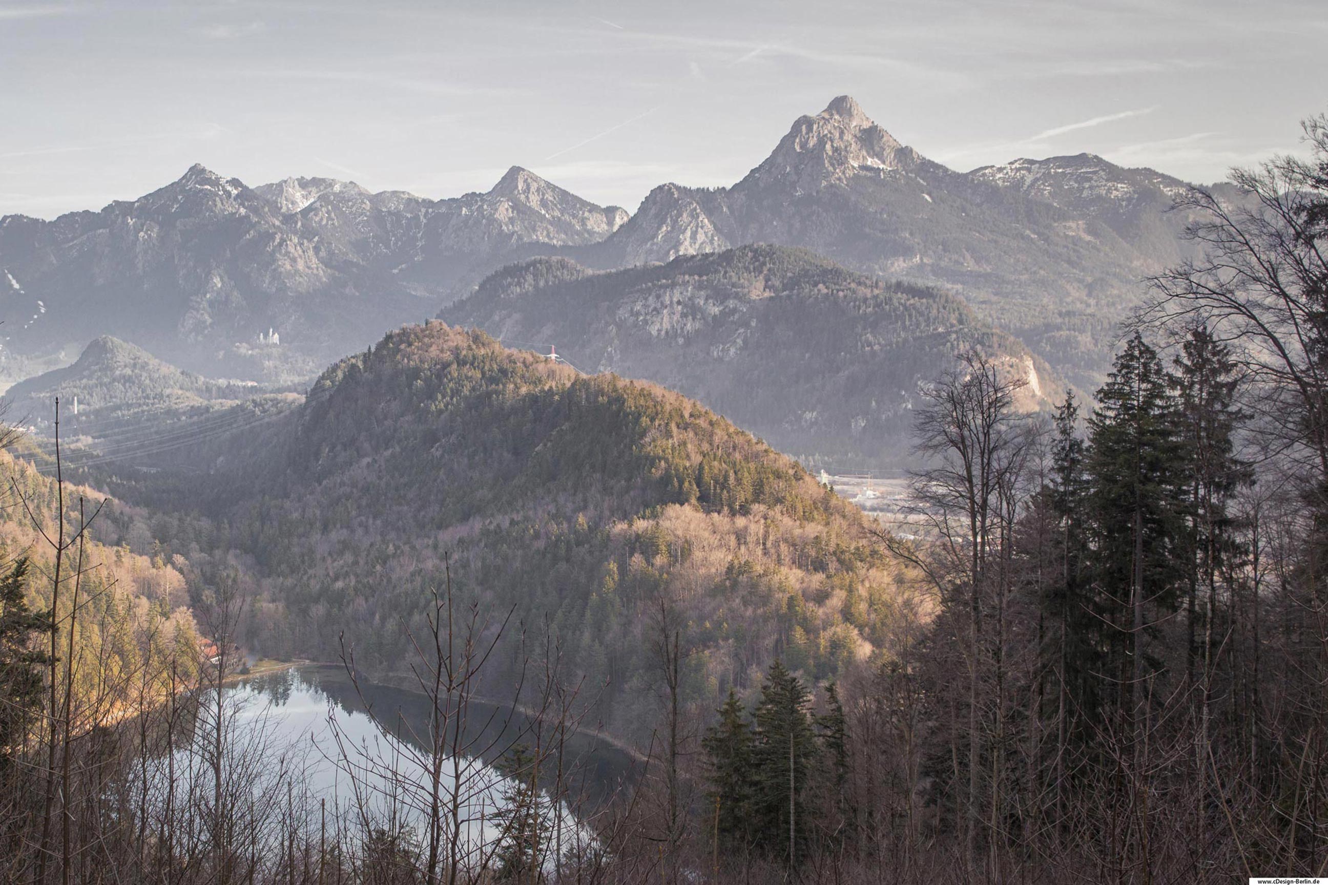Ich befinde mich auf einem Berg und vor mir liegt der Alatsee. Dieser ist von Waldbergen umgeben. Der rechte Berg hat die Erscheinung eines Drachenrückens, der das kleine Haus am See beschützt. Hinter diesem Berg befinden sich weitere Berge, sowie die berühmte Zugspitze, der höchste Berg Deutschlands. Fernab des Bildes sieht man als Silhouette das Schloss Schwanstein. Am unteren Rand des Bildes sieht man einen Mischwald. Dieses Foto entstand in Österreich an einem Herbsttag.