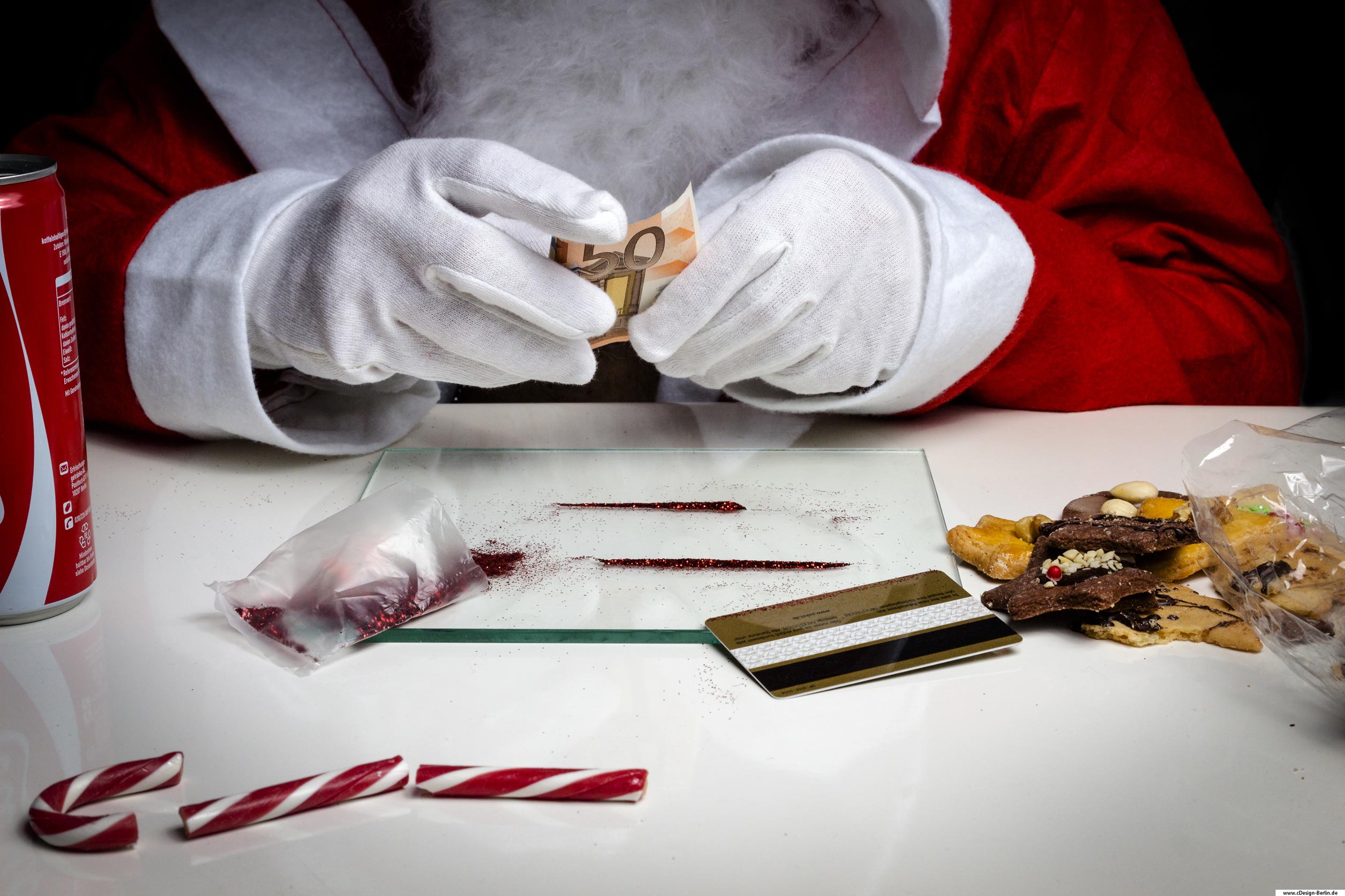 Oh du selige, oh du fröhliche Weihnachtszeit. Wie weit sind wir von dem eigentlichen Weihnachtsfest entfernt und hat es in unserer heutigen Zeit überhaupt noch einen Stellenwert? Wie wichtig ist uns das besinnliche Fest im Kreise unserer Liebsten? Wir fallen dem Konsumrausch zum Opfer und sind süchtig nach mehr! Auf diesem Foto sieht man einen Weihnachtsmann, der einen Geldschein über dem Tisch rollt. Auf dem Tisch befindet sich eine kleine Glasplatte, Glitzerstaub zu einer Line gezogen. Am linken Rand der Glasplatte eine Tüte mit rotem Glitzer und rechts eine Ec- Karte. Neben der Glasplatte rechts selbstgebackene Plätzchen und links eine Dose mit koffeinhaltigem Getränk. Am vorderen Bildrand liegt eine in drei Teilen zerbrochene Zuckerstange.