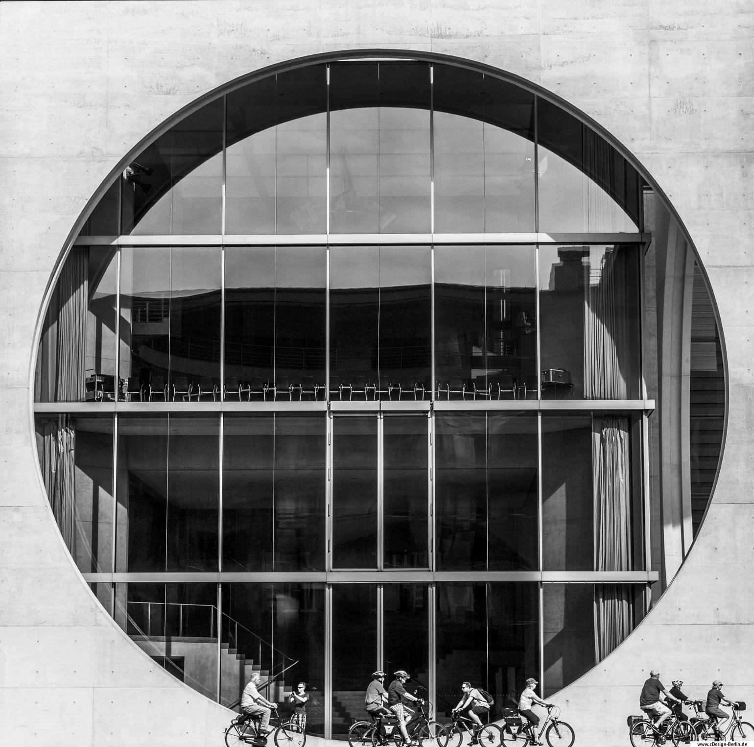 Das Bundeskanzleramt ist nicht nur der Sitz/ Büro unserer Bundeskanzlerin Angela Merkel, sondern auch eine Sehenwürdigkeit. Auf Grund des architektonischen Gebäudes, zieht es viele Touristen aus aller Welt an. Von den Berliner Bürgern liebevoll die Waschmaschine genannt, da dieses Gebäude im hinteren Teil sehr stark an eine Solche erinnert. Auf diesem Foto, sieht man in Nahaufnahme das große Auge der Waschmaschine. Vor diesem sind etliche vorbeifahrende Radfahrer zu sehen. Hierbei ist deutlich das Größenverhältnis zwischen dem Auge und der Radfahrer zu erkennen. Die Radfahrer wirken winzig klein, hingegen das Fenster übermäßig groß erscheint. Das Foto ist in schwarz/ weiß gehalten. Bezirk: Berlin- Mitte