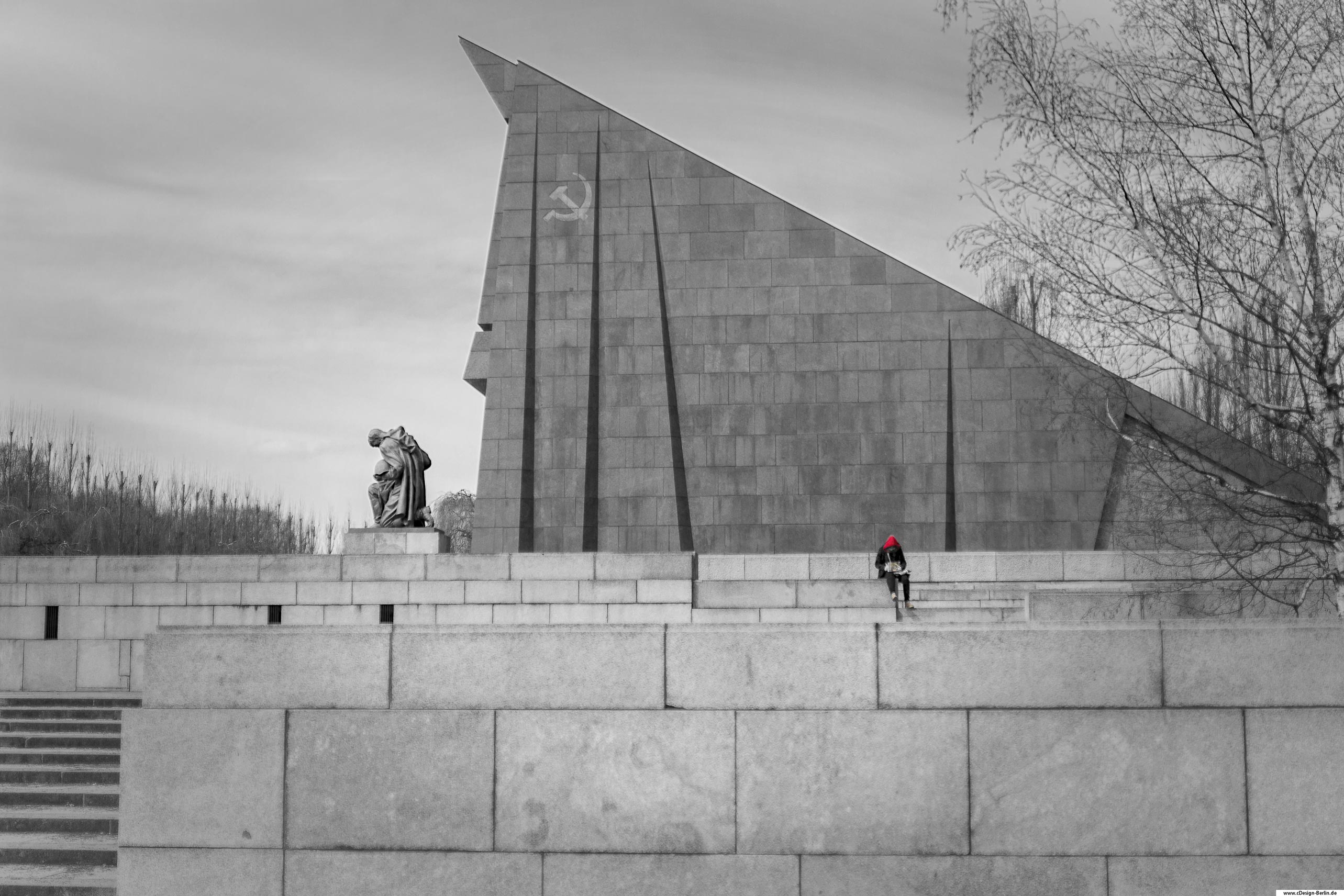Das sowjetische Ehrenmal im Treptower Park ist eine Gedenkstätte für die im zeiten Weltkrieg gefallenen Soldaten der roten Armee. Über 7000 von ihnen wurden hier bestattet. Der Bau des Denkmals stand im Zeichen des einsetzenden kalten Krieges. Mittig vom Bild ist die Statute eines Soldaten zu sehen, seinen Kopf gesenkt und in geknieter altung. Hinter ihm ein Monument, dessen Spitze in den Himmel ragt, auf diesem das Zeichen mit Sichel und Hammer zu sehen.Sein Helm liegend auf dem linken Oberschenkel. Er trägt einen langen Umhang. Ein paar Meter rechts von ihm sitzt eine Person auf einer Treppe, ihr Kopf ebenfalls gesenkt und von einer roten Kapuze bedeckt. Der Rest ihrer Kleidung schwarz. Der rechte Bildrand von einer Birke umrahmt. Foto schwarz/ weiß Kapuze rot Bezirk: Berlin-Treptow