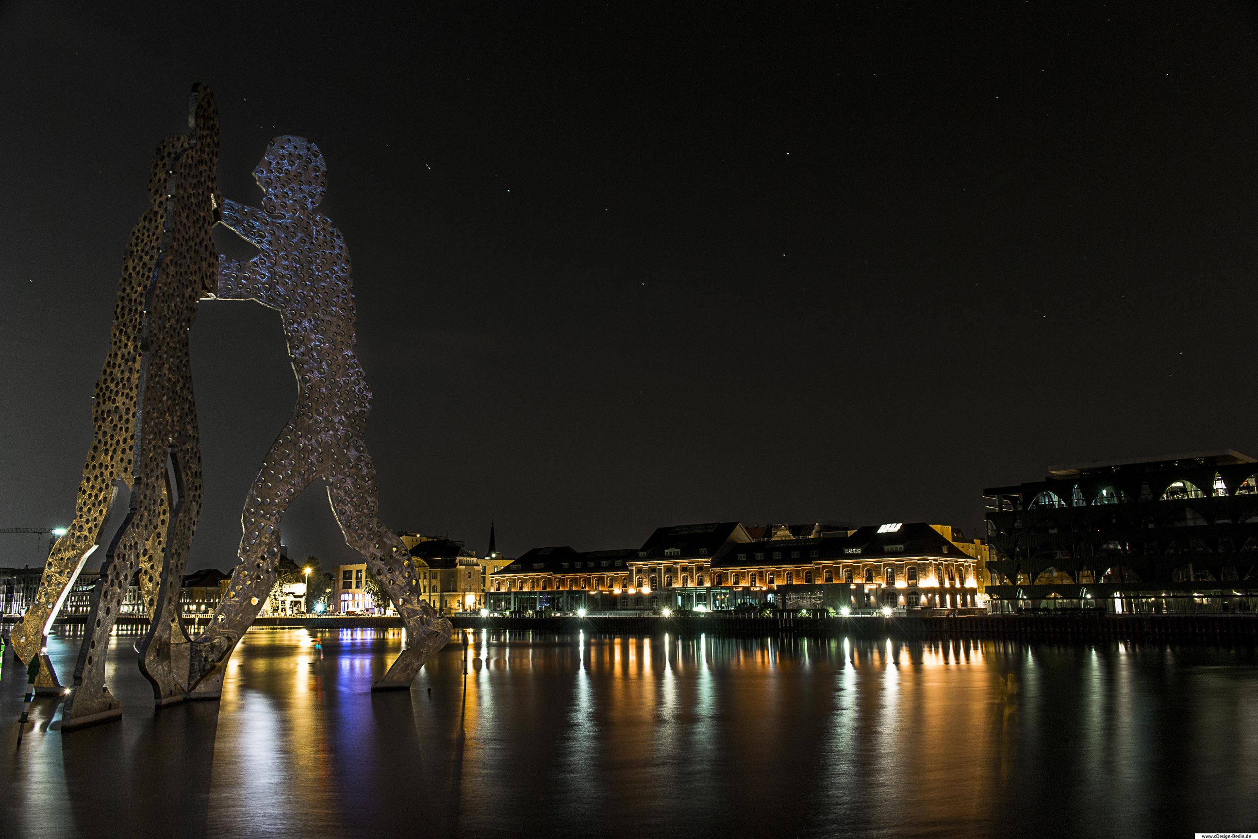 Die Skulptur soll daran erinnern, dass sowohl der Mensch als auch die Moleküle in einer Welt der Wahrscheinlichkeit existieren und es das Ziel aller kreativen und geistigen Tradition ist, Ganzheit und Einheit innerhalb der Welt zu finden. Zitat: Jonathan Borofsky Hierbei handelt es sich um eine Dreifachfigur. Sie ist die Nahtstelle des widervereinigten West-und Ostberlin und verbindet die drei vorherigen selbstständigen Berliner Bezirke Friedrichshain, Kreuzberg und Treptow. Am linken Bildrand befinden sich die Molekülmännchen inmitten der Spree. Deutlich ihre mit Löchern versehenen Körper zu sehen. Am rechten Bildrand, hintergründig Gebäude abgebildet. Das Foto ist in einer Sommernacht entstanden. Bezirk: Berlin- Friedrichshain, Kreuzberg, Treptow