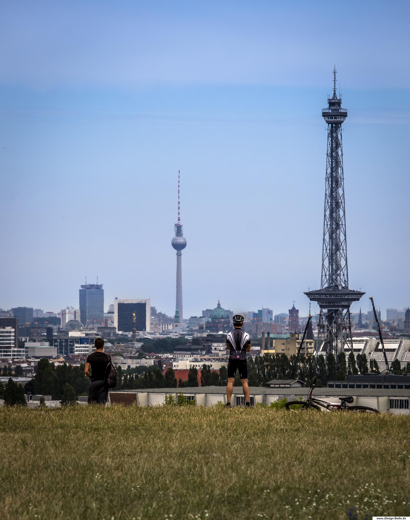Der Drachenberg ist eine hohe Erhebung im Bezirk Charlottenburg- Wilmersdorf in Berlin. Er liegt im Ortsteil Grunewald. Man nennt ihn auch den kleinen Teufelsberg. Der Drachenberg entstand nach dem Zweiten Weltkrieg aus Trümmerschutt. Dieser ist Begegnungsstätte der Berliner/innen und lädt zum entspannen, plaudern und diversen sportlichen Aktivitäten ein. Auch die Herzen der Fotografen, sowie Hobbyfotografen schlagen höher, bei einer wundervollen Aussicht über Berlin. Im Vordergrund des Bildes ist eine Wiese zu sehen und am Bergrand stehen rechts ein Radfahrer, sein Fahrrad im Gras liegend und links vom Bild ein weiterer Mann. Beide genießen den wunderbaren Ausblick, denn vor ihnen ist rechts im Bild der Funkturm zu sehen und am Horizont der Fernsehturm. Die Aussicht ist klar und hätte nicht besser sein können, am diesem Sommertag. Bezirk: Berlin Charlottenburg-Wilmersdorf, Ortsteil Grunewald