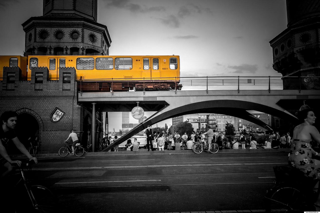 Die Oberbaumbrücke ist das Wahrzeichen des Bezirkes Friedrichshain-Kreuzberg. Sie verbindet als Teil des Innenstadtrings die Ortsteile über die Spree und liegt zwischen der Elsenbrücke und der Schillingbrücke. Für die Touristen unserer Stadt ein beliebter Anziehungspunkt und gern überfüllt. Ausserdem ist dieser Ort für seine Vielzahl von Radfahrern bekannt. Am vorderen Bildrand sind rechts und links Fahrradfahrer abgebildet. Der linke Radfahrer sportlich, in Eile und die rechte Radfahrerin entspannt und gelassen, in einem Sommerkleid mit Blumenmuster. Mittig vom Bild direkt vor der Brücke befinden sich zwei weitere Radfahrer mit Abstand. Der linke Radfahrer, seinen Kopf über die rechte Schulter gedreht, kommuniziert mit einer weiteren Person die sich mittig zwischen den Radfahrern befindet. Der rechte Radfahrer seinen Blick auf eine Ansammlung von Menschen gerichtet, dessen Aufmerksamkeit den Musikanten gewidmet wurden. Auf der Brücke fährt gerade die U-Bahn ein. Hinter der Brücke links und rechts die Türmchen zu sehen. Foto in schwarz/ weiß. Bezirk: Berlin- Friedrichshain