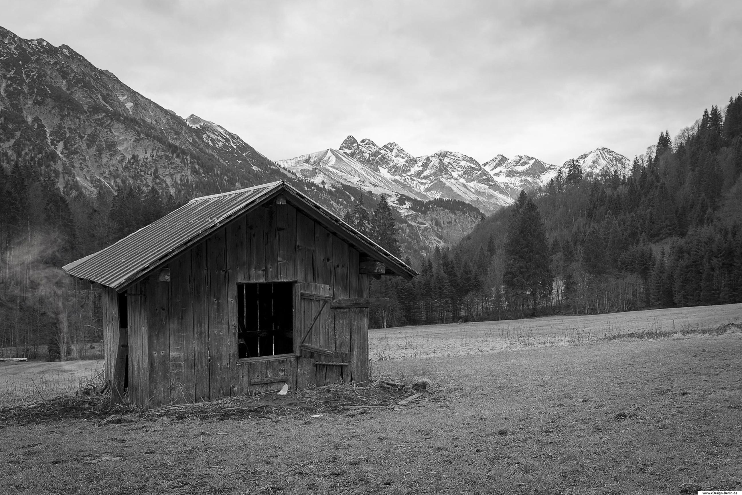 Es ist kalt und ich befinde mich in den Bergen auf einer Wiese. Vor mir steht eine kleine Scheune, die ihre Tage schon gezählt hat. Ihr Dach ein altes Wellenblech. Das Fenster der Scheune steht offen, sie lädt zum hineingehen ein. Hinter ihr große Berge deren Spitzen mit von Schnee bedeckt sind. Rechts von der Scheune ein paar Meter weiter kommt man in eine Waldlichtung. Es ist ein kalter und Wolkenverhagener Tag, deshalb habe ich dieses Foto in schwarz / weiß gehalten.