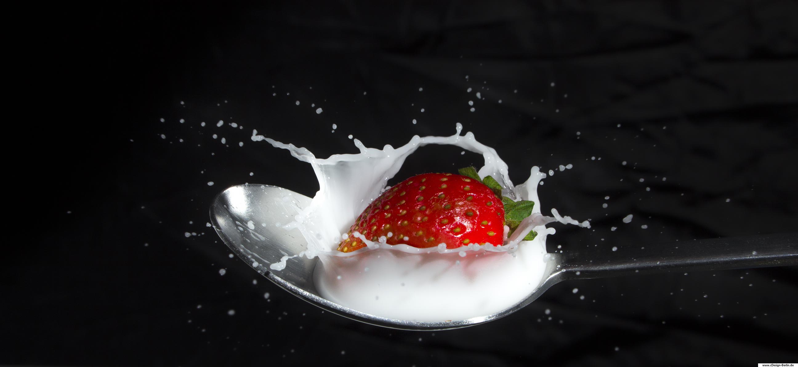 Es ist heiß und du möchtest dich in Form eines Getränkes oder Obstes erfrischen. Auf diesem Foto sieht man einen Edelstahhllöffel der mit Milch gefüllt wurde. Eine Erdbeere fällt in die Milch hinein und es entsteht ein wundervolles Zusammenspiel zwischen diesen beiden Elementen. Die Milch erzeugt eine Welle die sich in alle Richtungen ausbreitet, wenn die Erdbeere auf diese trifft. Die Welle der Milch wirkt eingefroren, da die Verschlusszeit beim Auslösen kurz gehalten wurde. Der Hintergrund auf diesem Foto ist schwarz gehalten, somit kommt das Zusammenspiel dieser Elemente sehr gut zum Tragen. Wie wäre es? Lust auf Erdbeermilch?