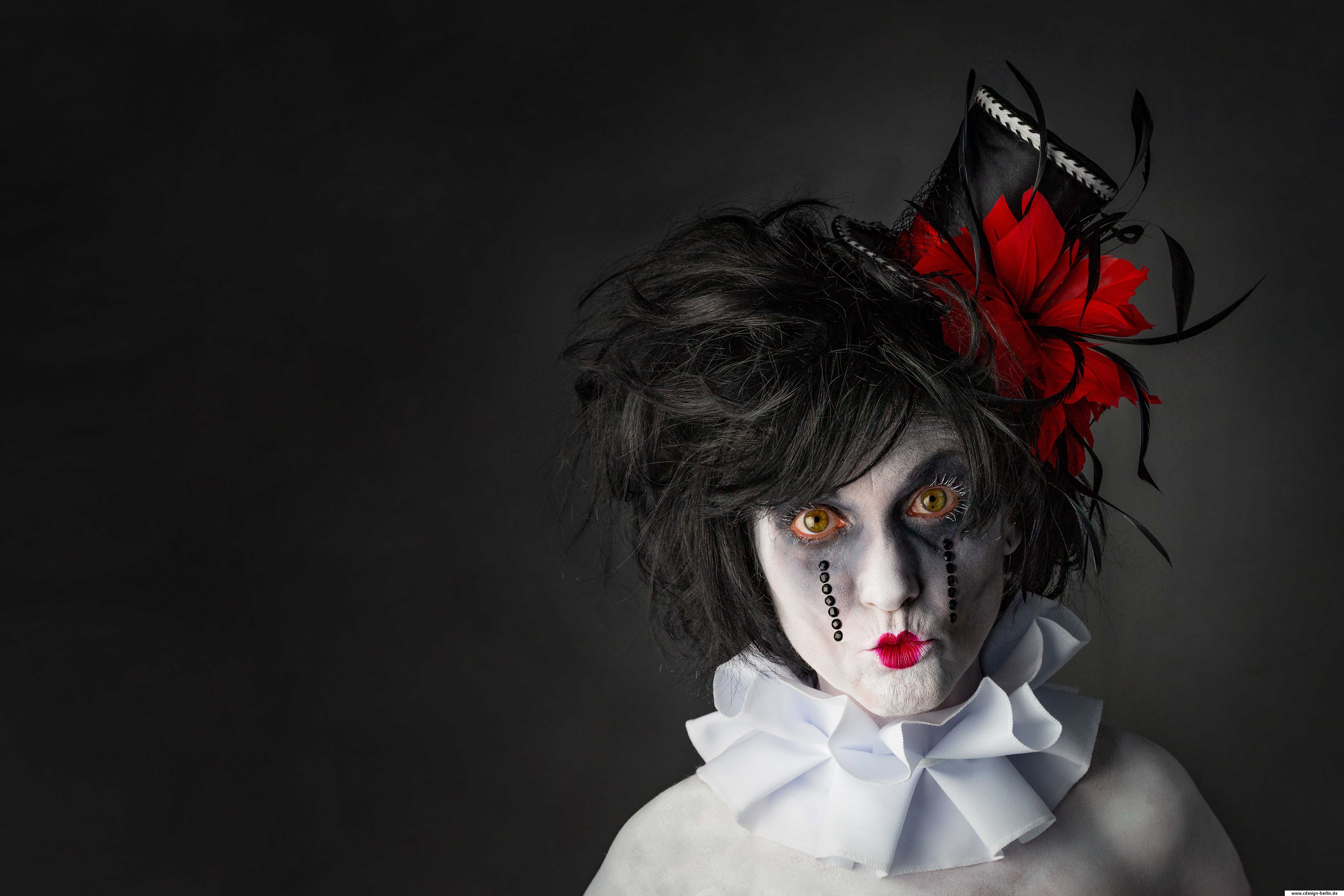 Dein Aussehen ist nur eine Hülle und dein Inneres bleibt verborgen. Wir geben gern den Clown um nicht hinterfragt zu werden. Wie nehme ich eine Maske ab, wenn sie auhört eine Maske zu sein, wenn sie so sehr ein Teil von mir ist wie ich Selbst. Wie ein Clown aussieht bedarf nicht vieler Worte, da sie sich im Laufe der Zeit nur unwesentlich in ihrer Darstellung geändert haben. Auch auf diesem Bild gibt es keine wesentlichen Veränderungen, der einzige Unterschied liegt darin, keine rote Nase vorzufinden. Die Augenränder des Models sind dunkel gehalten, der Mund gespitzt, um dadurch eine Herzform zu erhalten. Ihre Haare sind zersaust, schwarz und mit einem Hut verziert. Der Hut ähnelt einem Zylinder ( Miniaturausgabe), verziert mit einem Band in einem knalligen Rotton. Das Band ist seitlich mit großen roten Federn versehen, die der Form einer Blüte sehr nahe kommt. Im Blütenkern erstrecken sich weitere Federn, lang und schmal gehalten. Diese haben widerrum Ähnlichkeit mit Vogelfedern eines Raben. Um den Hals trägt das Model eine große weiße Krempe, die an alte Barockzeiten erinnert. Ihr Blick ist leer und zerbrechlich.
