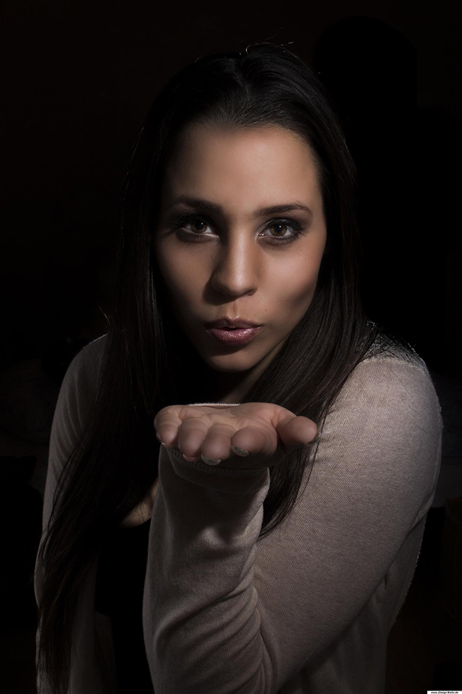 Ein Luftkuss ist wie ein warmer Sommerregen. Auf diesem Foto ist eine junge Frau zu sehen, die gerade einen Luftkuss verschenkt. Der Hintergrund ist schwarz gehalten, somit fällt der Blick des Betrachters sofort auf das Gesicht. Ihre Haut ist glatt, seidig schimmernd und weich gehalten. Ihre Haare sind dunkelbraun und lang. Ihre braunen Augen strahlen Wärme aus und tragen ein Lächeln in sich.