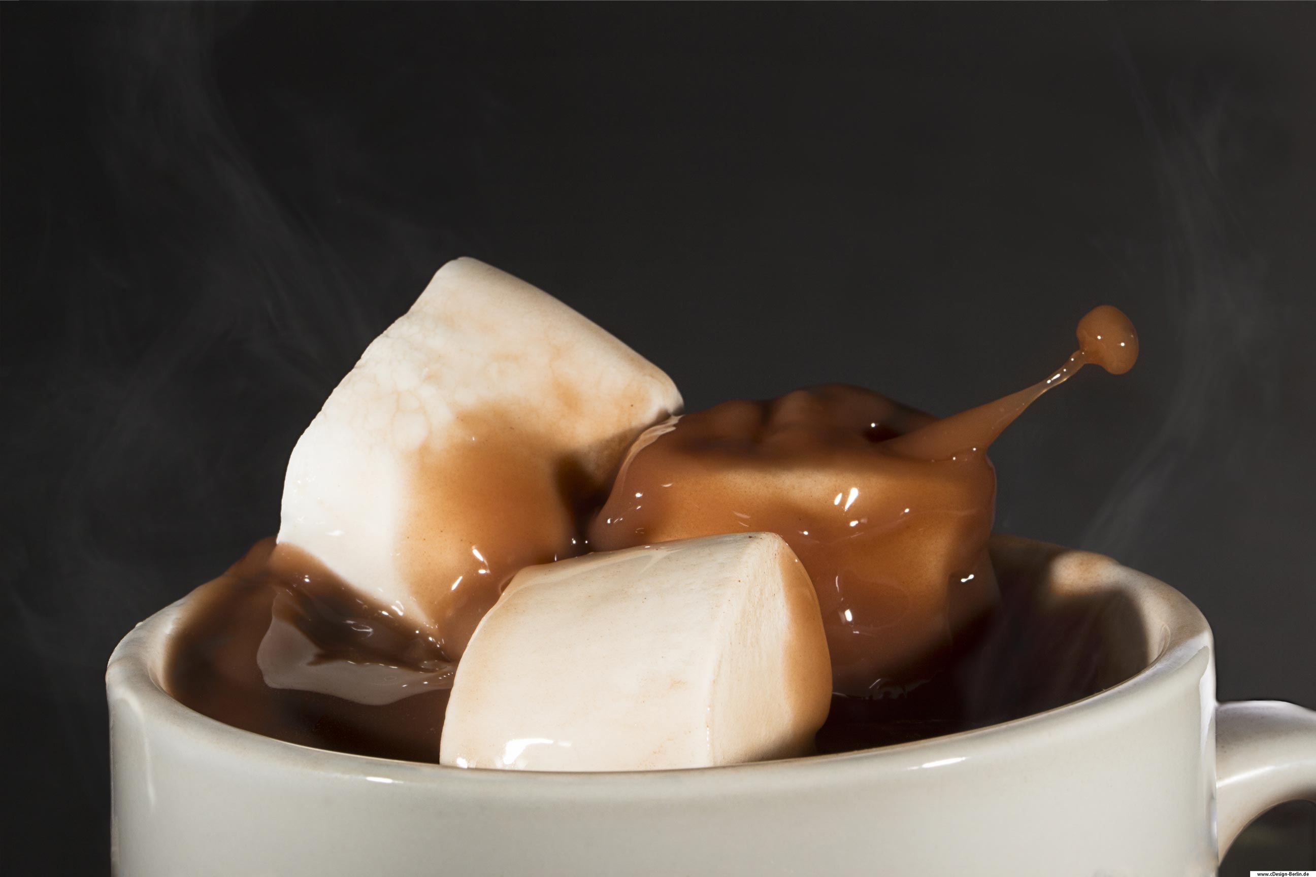 Du verspürst Heißhunger auf etwas Süßes. Es ist Winter und du möchtest es dir zu Hause auf deinem Sofa gemütlich machen. Wie wäre es mit einer heißen Schokolade und Marshmallows darin? Hier siehst du eine Tasse mit heißer Schokolade. Die Marshmallows fallen sanft hinein und verursachen ein überschwappen der Trinkschokolade. Dampf steigt aus der Tasse empor. So klingt ein kalter Winterabend aus.