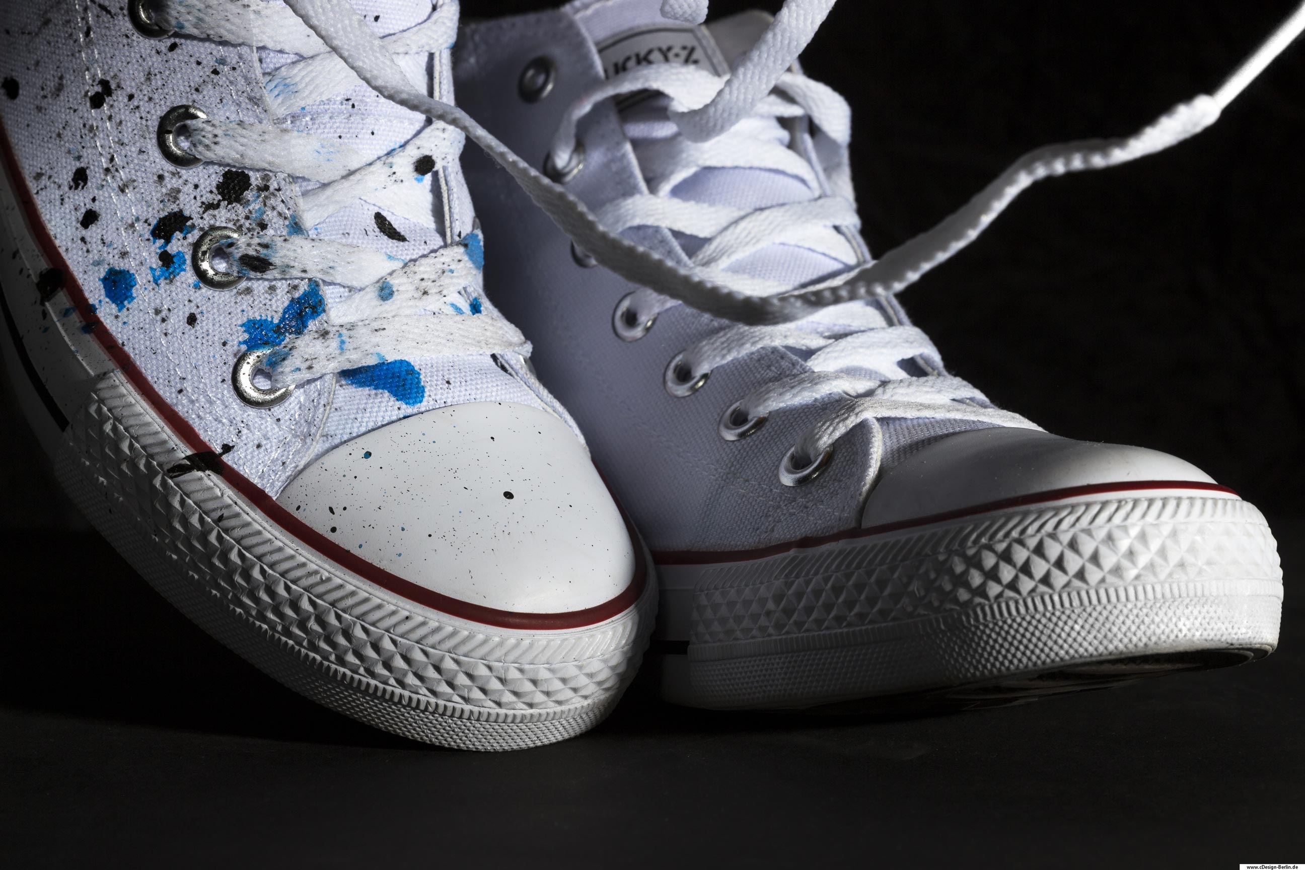 Möchtest du Tanzen, dann schlüpfe in deine Schuhe und fühle den Beat. Der Hintergrund dieses Fotos ist in schwarz gehalten und gibt einen guten Kontrast zu den weißen Converse Schuhen. Die Schuhe wurden lebendig aufgestellt, der Linke steht und bei dem rechten ist die Hacke erhöht, wie beim Gang. Der linke Schuh weist noch eine entscheidende Besonderheit auf. Er wurde durch eine Künstlerin mit azurblauen und schwarzen Farbkleksen versehen. Der Schnürsenkel des linken Schuhs scheint in der Luft zu schweben.