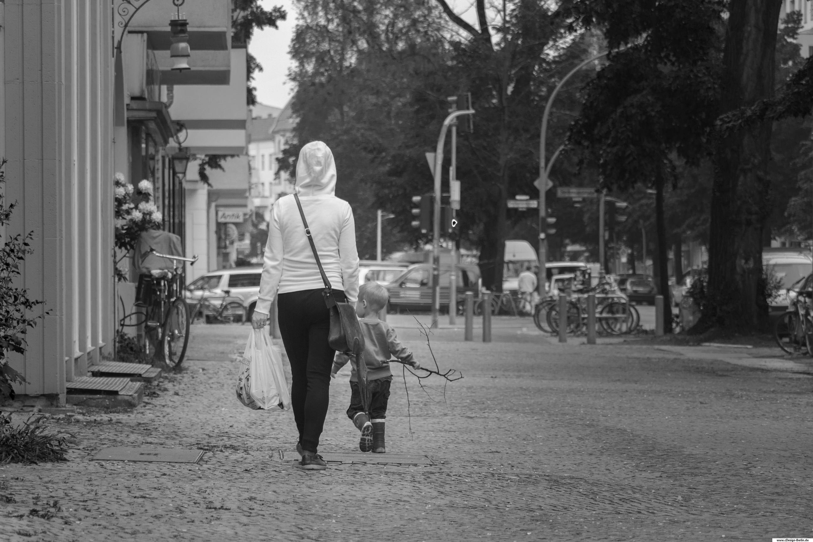 inder entdecken auf ihre ganz eigene Art die Welt und deren Umgebung. Ein Einkauf mit der Mutter kann zu einem kleinen Abenteuer werden, um nicht der langen Weile zum Opfer zu fallen.
Erwachsenensachen sind für Kinder nicht greifbar, eher lanweilig, weil es nicht ihrem eigenen Kosmus entspricht. Auf diesem Foto sieht man eine Mutter mit ihrem Sohn. Sie trägt eine Tüte in ihrer linken Hand mit Dingen die sie eingekauft hat. Eine Umhängetasche rundet ihr Gesamterscheinungsbild ab. Ihr Sohn läuft einen Meter vor ihr, in seiner rechten Hand einen Stock haltend. Der Blick des Jungen richtet sich nach links eines Wohnhauses, vielleicht hat er etwas Spannendes entdeckt. Die Mutter hingegen ihren Blick nach vorn gerichtet, der Kopf von der Kapuze ihrer Jacke bedeckt. Links ein paar Meter weiter steht ein Fahhrad mit Kindersitz. Über dem Fahhrad hängen große Blüten eines Busches, fast einem Gemälde gleichend. Mutter und Kind steuern auf eine Strasse zu. Rechts von ihnen die große Hauptstraße. Dieses Bild strahlt für mich eine unglaubliche Ruhe aus, als ob die Zeit für einen Moment lang angehalten wurde, denn außer den beiden weit und breit keine Menschenseele. Auch dieses Foto habe ich in schwarz/ weiß gehalten.