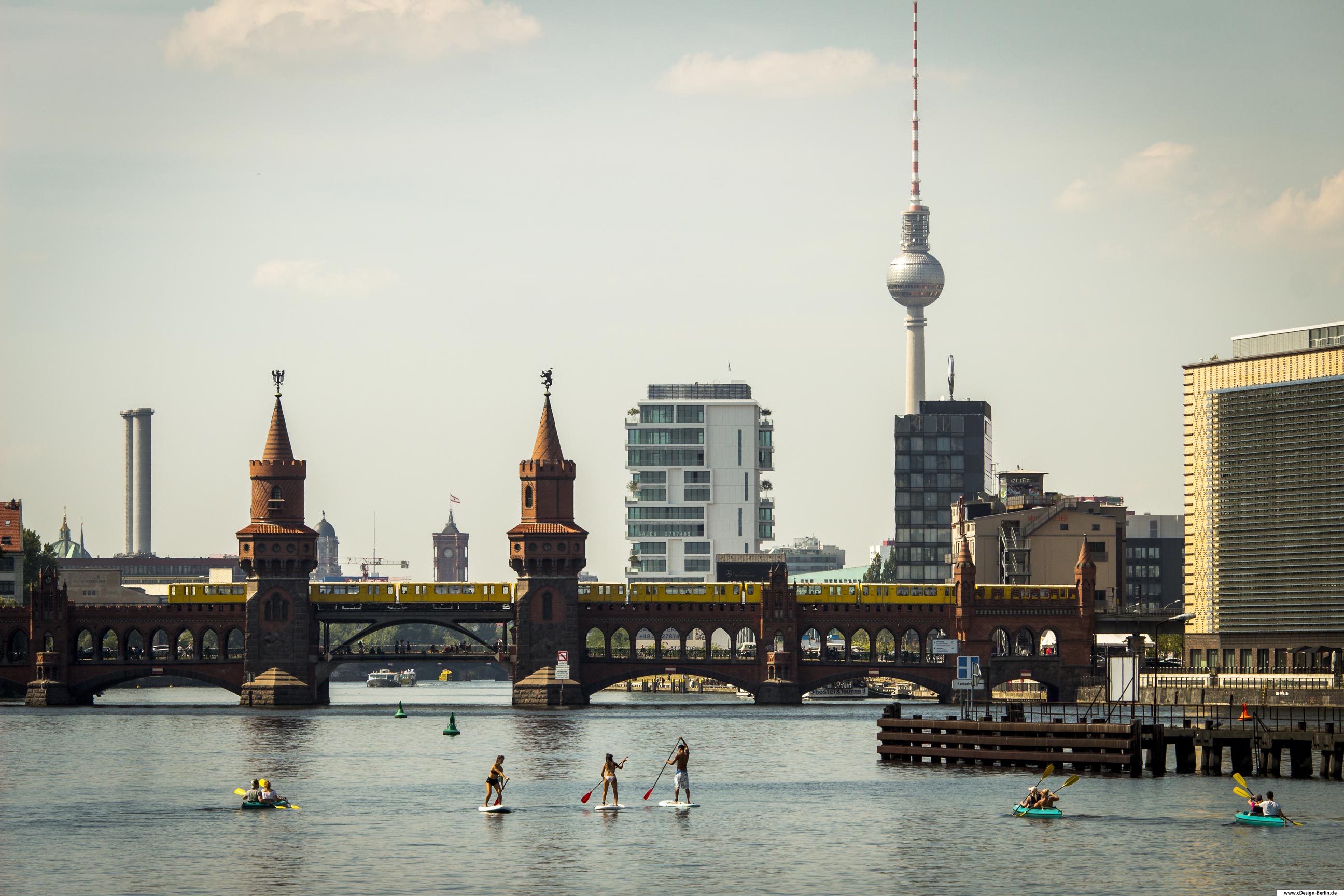 Im Sommer zieht es viele Touristen nach Berlin. Ein beliebter Ort ist die Oberbaumbrücke mit ihren kleinen Türmen. Hier tummeln sich Menschen aller Herrenländer. Die Spree fließt unter der Brücke hindurch. Nicht weit davon befindet sich der Alexanderplatz mit seinem Wahrzeichen, dem Fersehturm. Dieses Foto zeigt die Oberbaumbrücke, auf ihr die U- Bahn fahrend. Rechts vom Bild befindet sich ein Kastenförmiges Gebäude. Hinter der Brücke sind weitere Gebäude, die Dachspitze des roten Rathauses, sowie der Fernsehturm zu sehen.
Auf der Spree genießen Kanufahrer und Paddelsurfer den herrlichen Sommertag.
